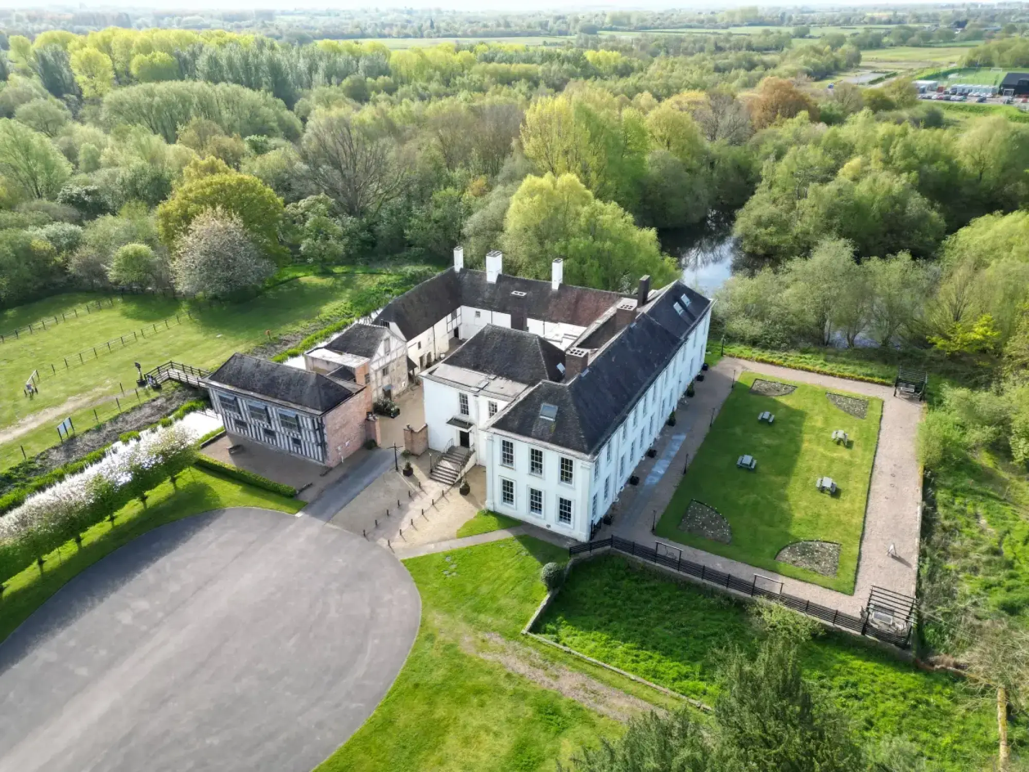 An aerial view shows a large, U-shaped historic hall with white rendered walls and a timber-framed wing, surrounded by formal lawns, rectangular planting beds, mature trees, and a winding river.