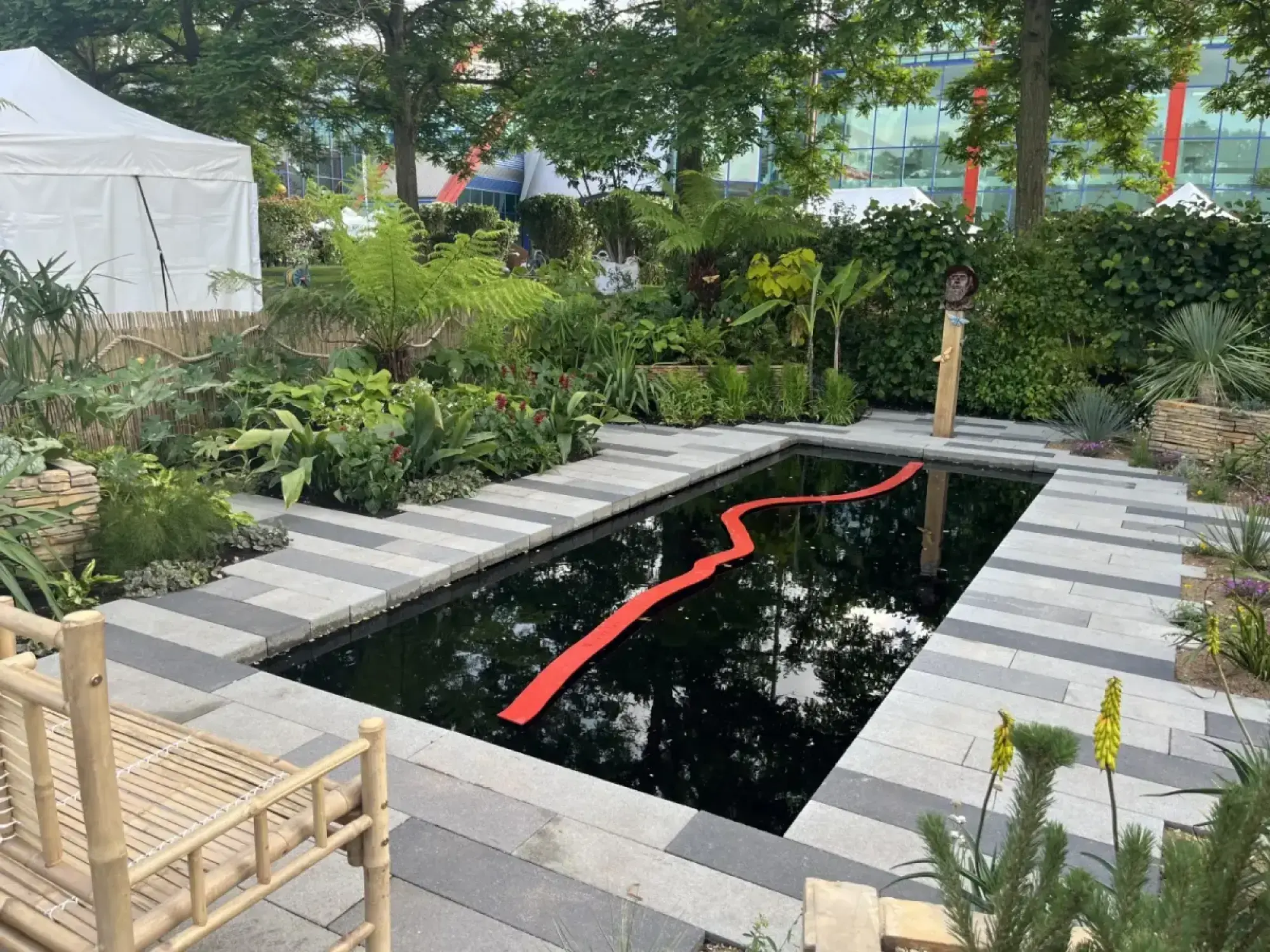 A contemporary garden with a black rectangular pond featuring a wavy red line, bordered by lush tropical plants, a bamboo bench, and striped grey paving.