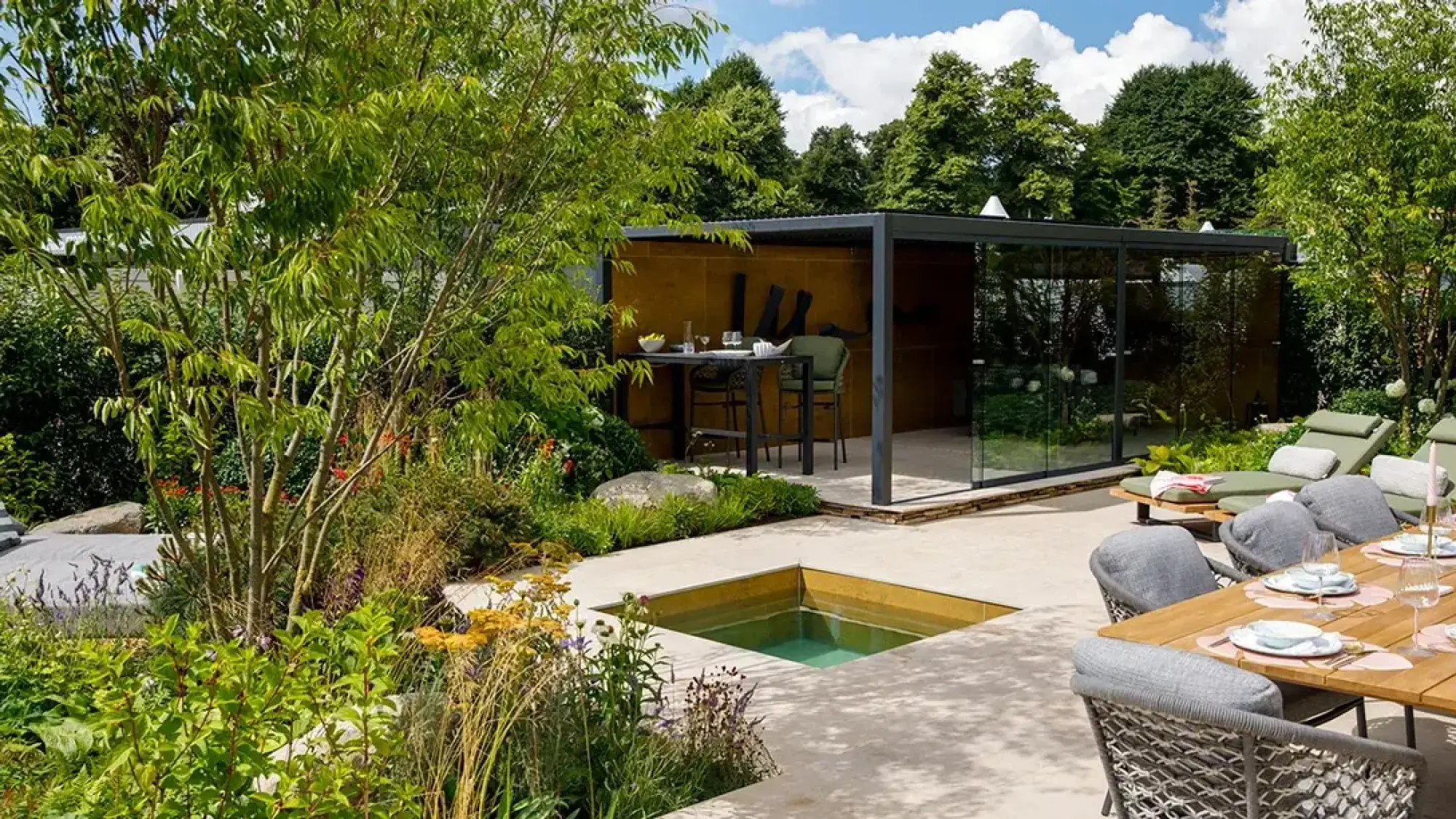A contemporary outdoor garden with a glass room, sunken pool, paved patio, dining set, sun loungers, and lush green border plants.