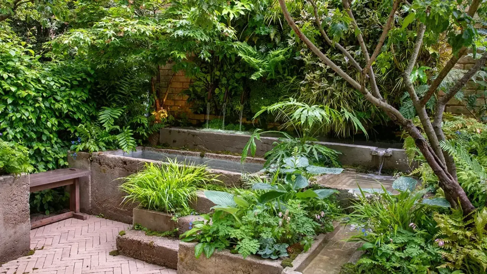 A tranquil container garden features tiered concrete planters with flowing water, lush green plants including ferns and hostas, a vertical green wall, and a wooden bench on a herringbone brick path.