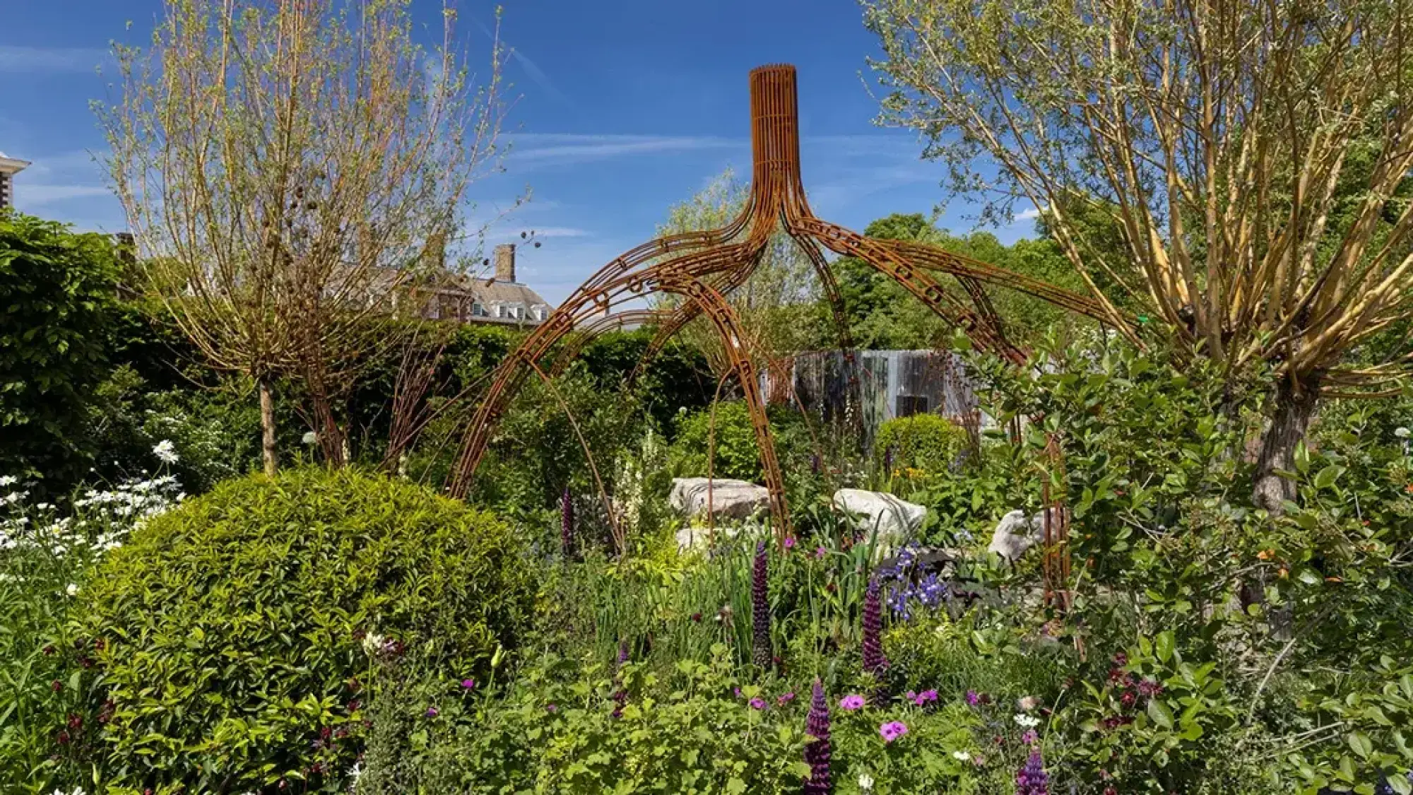 A lush, naturalistic show garden features a large, rusted metal sculpture resembling mangrove roots, surrounded by vibrant green foliage, purple lupins, and white daisies.
