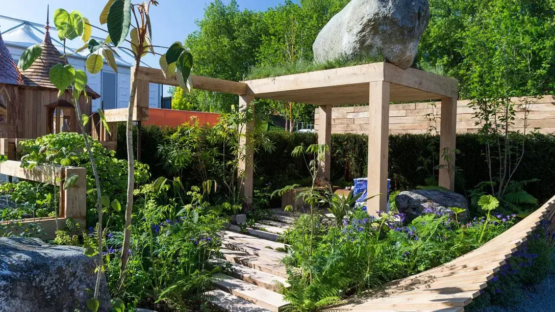 A fresh garden featuring a timber pergola supporting a large boulder and green roof, with a wooden playhouse and a winding path through lush plants.