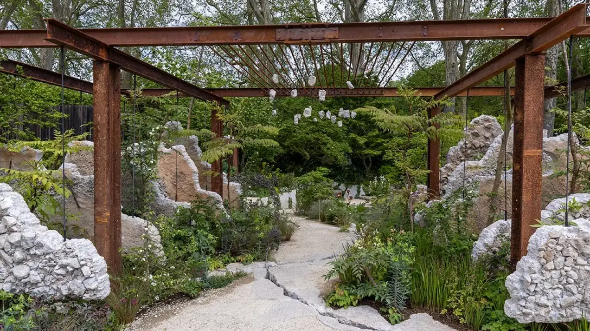 A show garden with a rusted steel pergola over a fractured path, surrounded by textured concrete rocks, lush green plants, and white hanging sculptures.