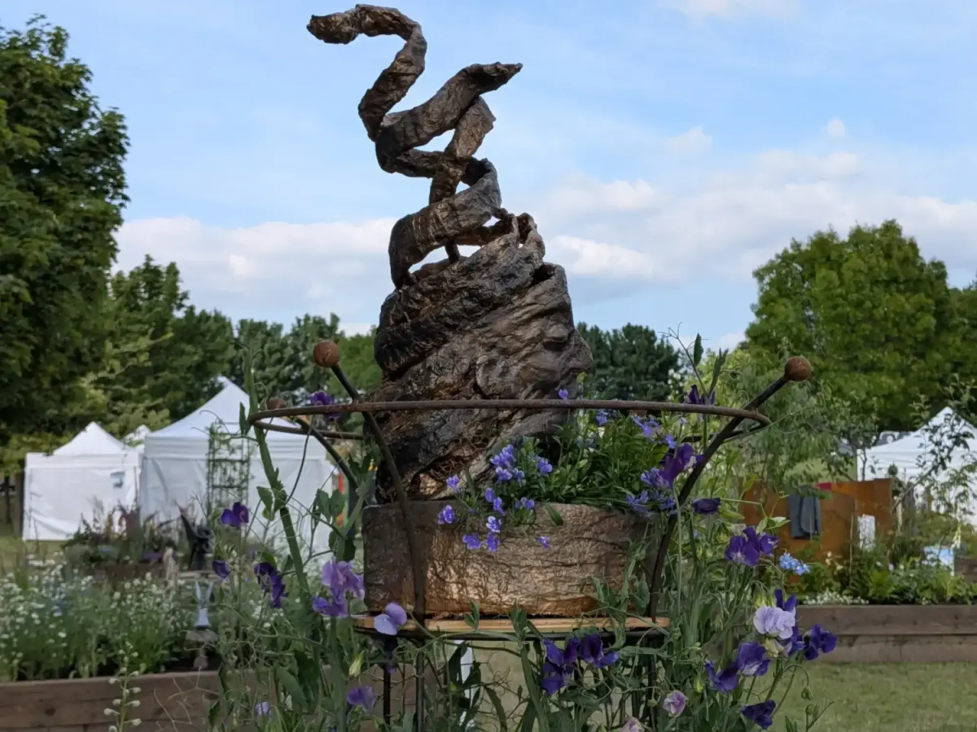 A bronze-effect spiral sculpture rises from a gold-leaf planter filled with purple sweet peas and blue flowers, set within a sunny border.