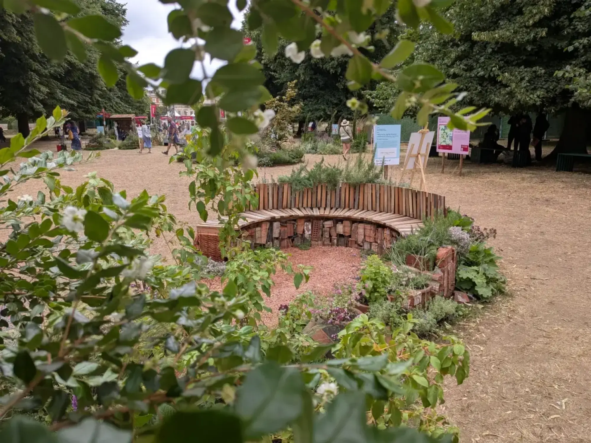 A semi-circular feature border with a curved wooden slatted bench backed by a low stacked brick wall, surrounding a reddish-brown mulch area. Mixed green planting with small purple flowers fills the foreground and sides.