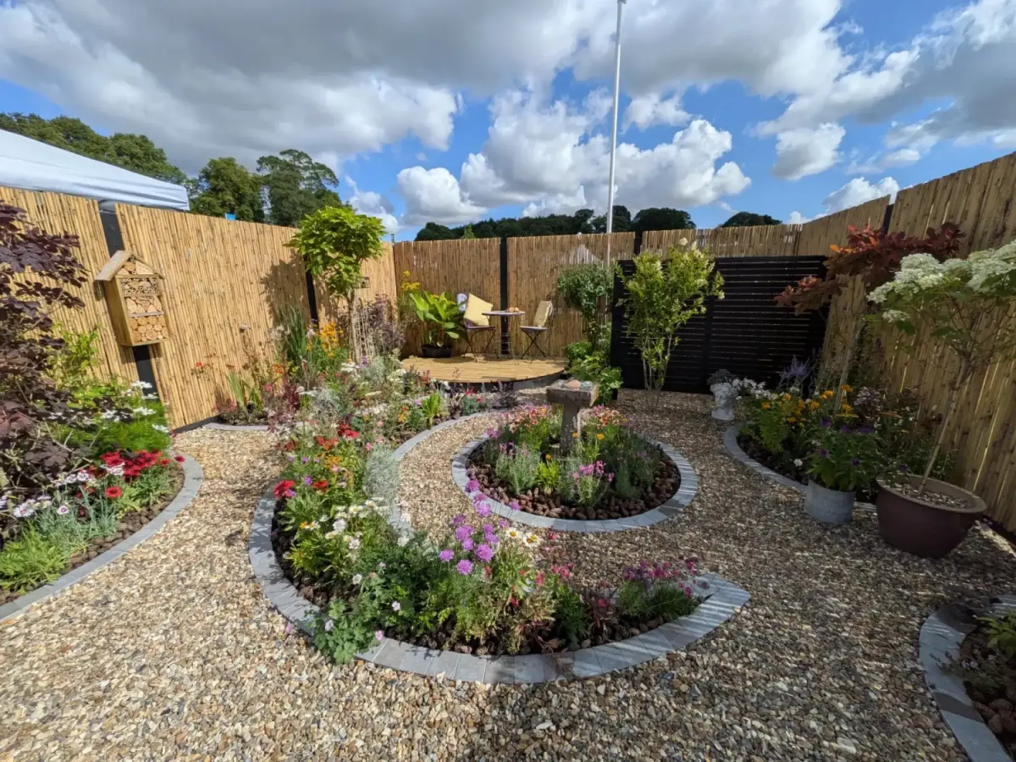 An inviting feature garden with curved gravel paths and raised beds filled with colourful flowering plants, a central birdbath, an insect hotel on a bamboo screen fence, and a wooden deck with a small table and chairs.