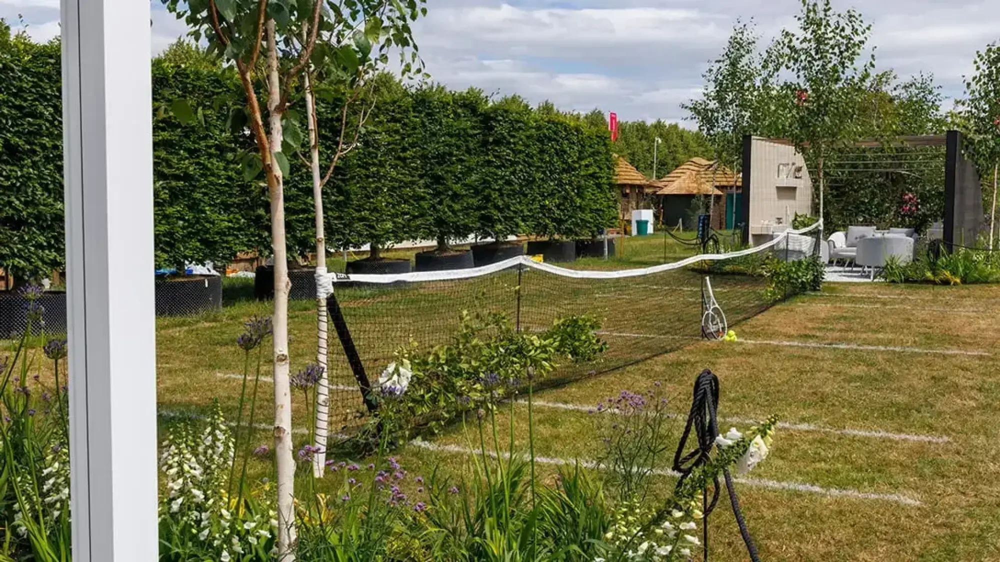 A modern garden with a grass tennis court and net, a grey outdoor lounge, dark pergola, silver birch trees, and white and purple flowers.