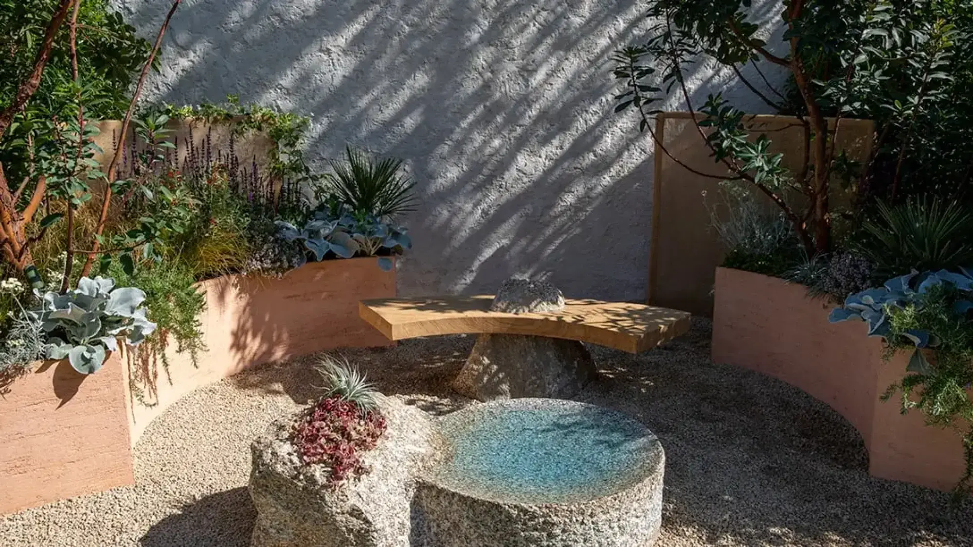 A tranquil Mediterranean container garden featuring a curved timber bench, a sculpted granite water feature, and terracotta-toned raised planters filled with drought-tolerant plants.