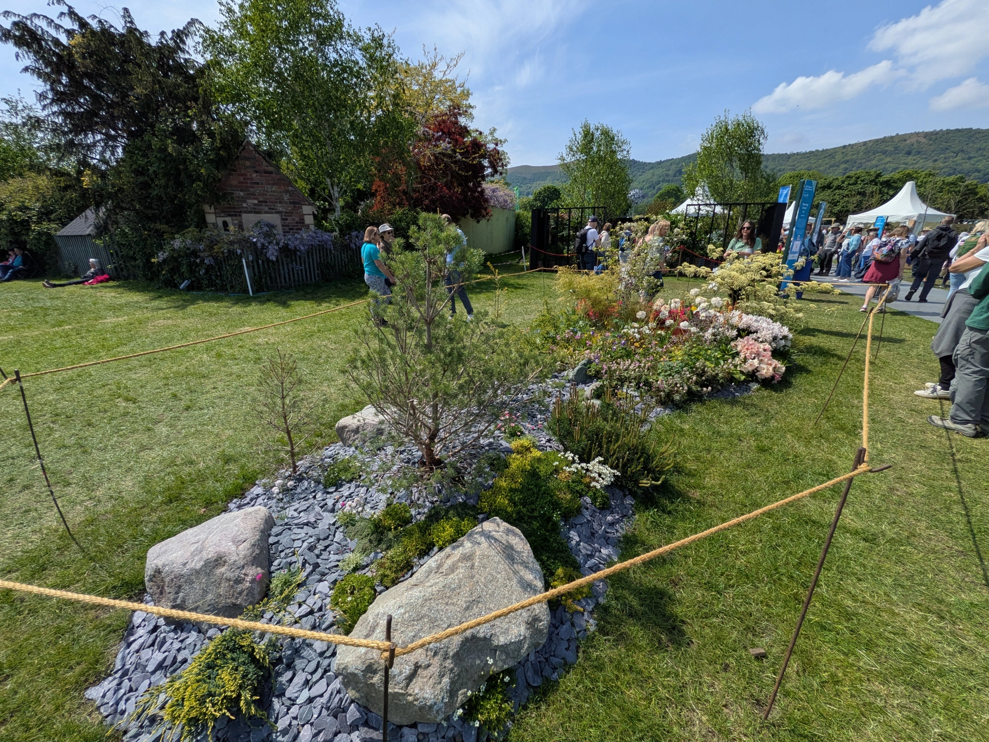 A beautiful border garden featuring large boulders, dark grey slate chippings, dwarf conifers, and a mix of green foliage and small white, pink, and purple flowers.