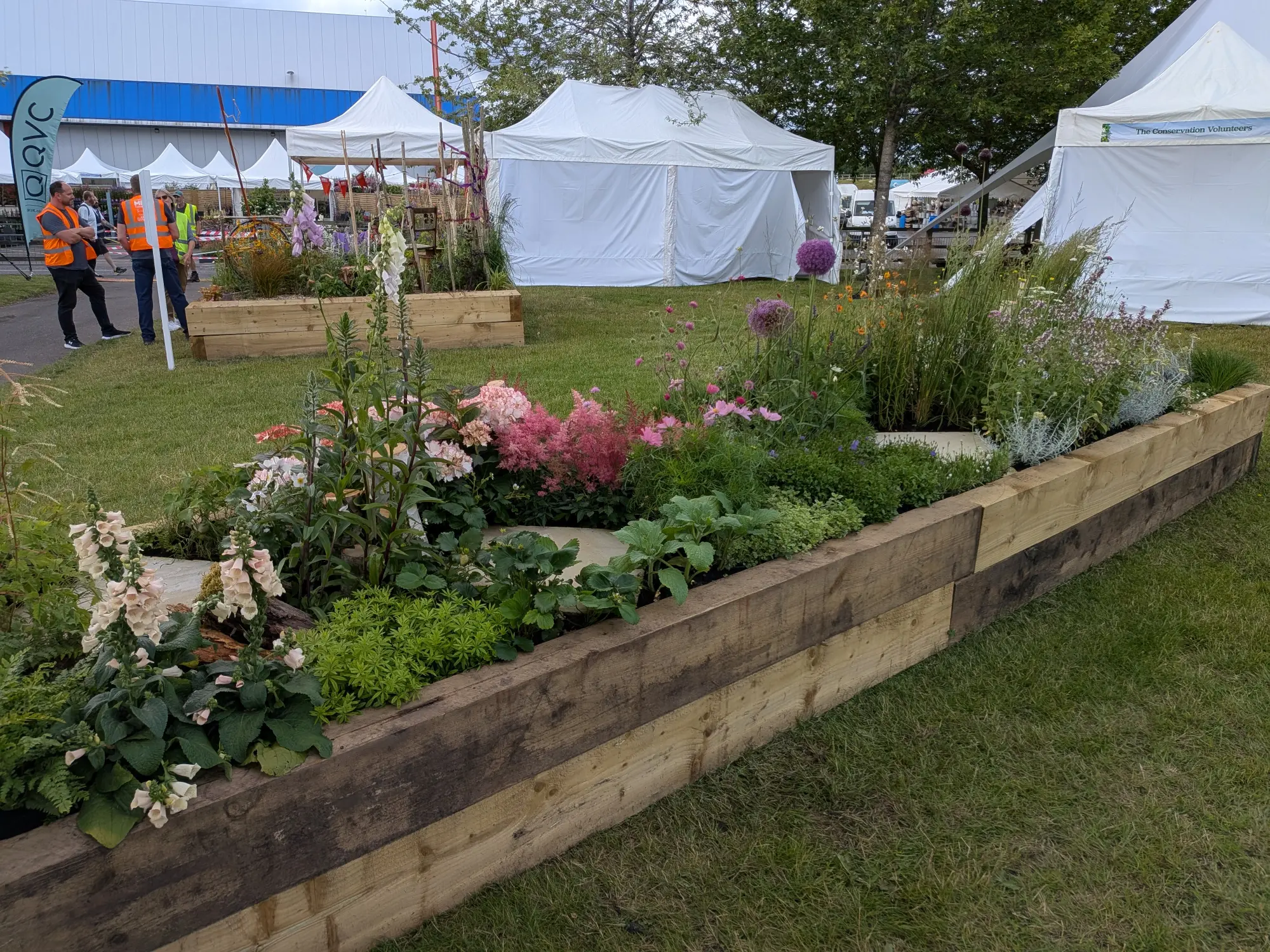 A long raised garden border made of stacked timber sleepers is filled with a lush, naturalistic planting of pink foxgloves, purple alliums, astilbe, and mixed perennials.