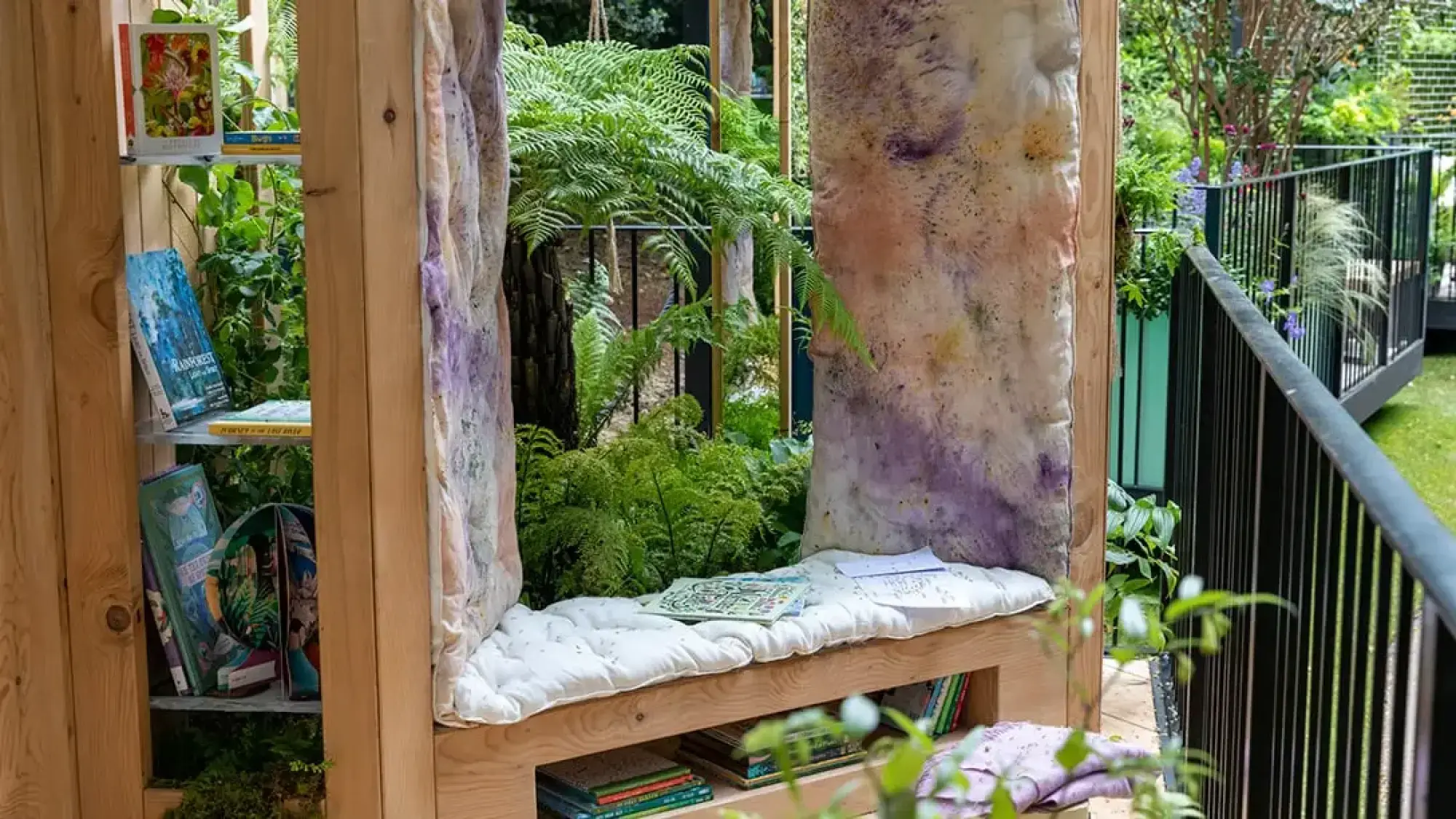 A cozy wooden reading nook on a balcony garden features built-in bookshelves, a cushioned bench, patterned fabric panels, and lush green ferns.