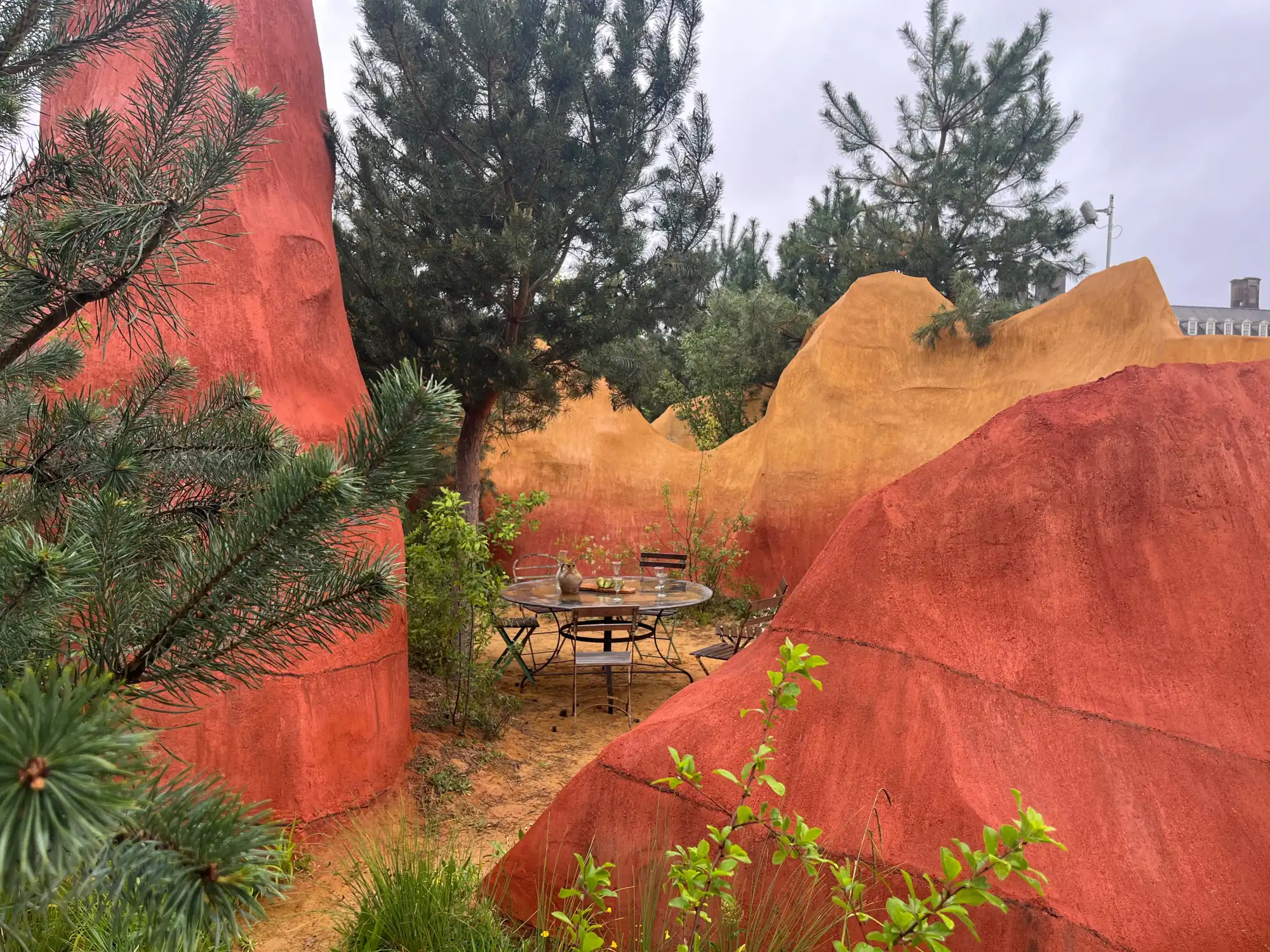 A sculptural garden features tall, undulating mounds in terracotta red and ochre orange, surrounding a metal dining table and chairs, with pine trees and sparse green plants.