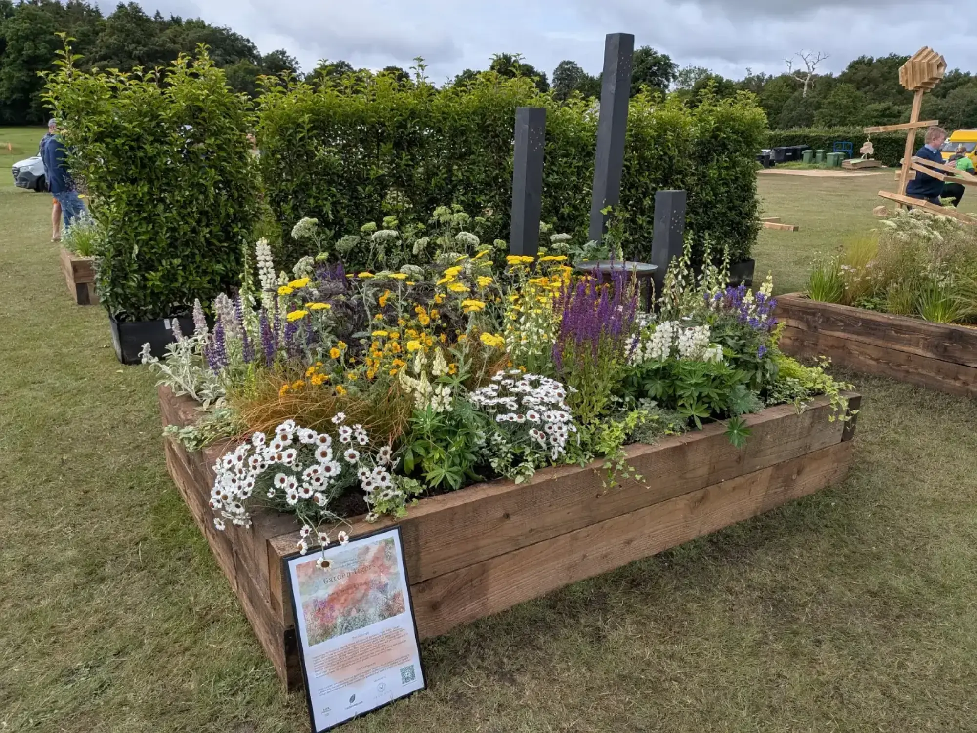 A vibrant show garden in a raised timber planter, with yellow, purple, and white perennial flowers, dark sculptural pillars, and a bird bath.