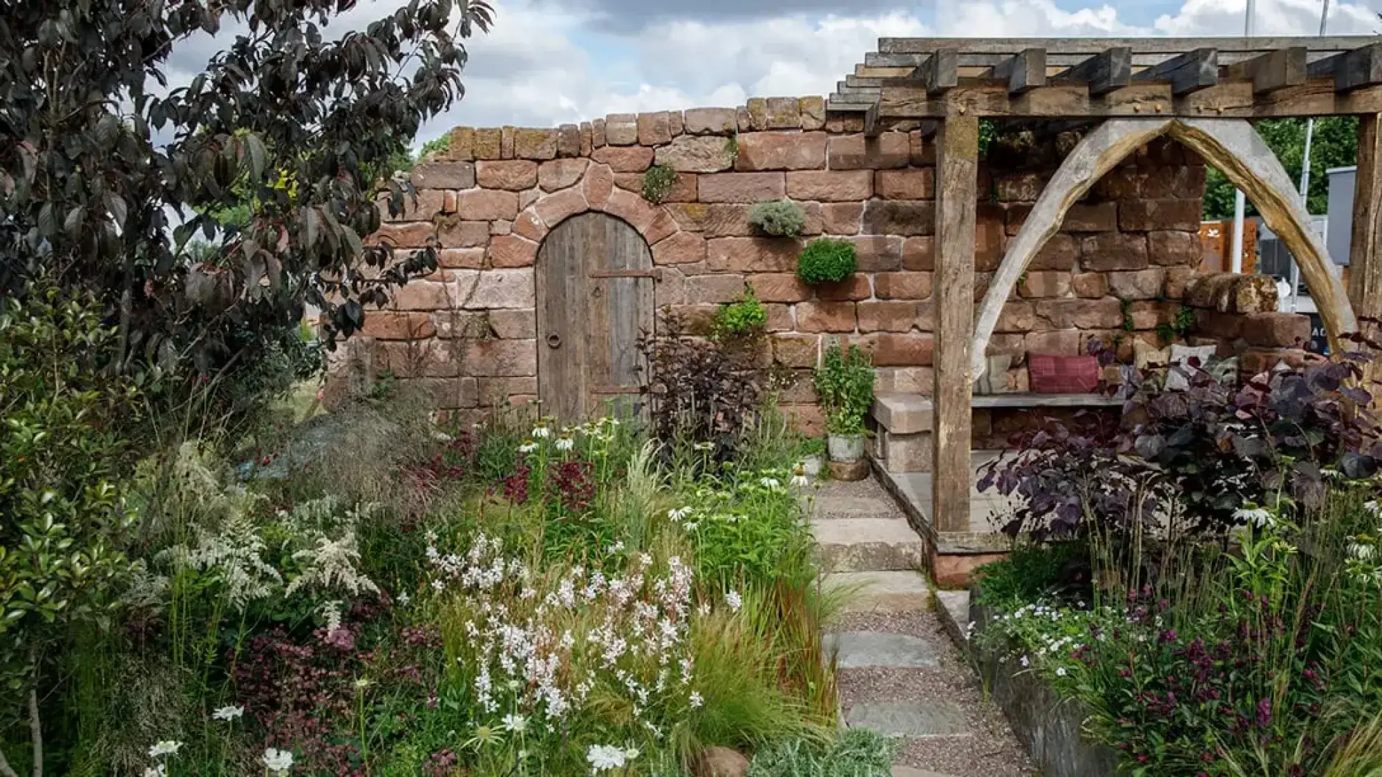 A naturalistic garden features a rustic stone wall, arched timber door, a weathered pergola, stone bench, and lush white and purple planting.