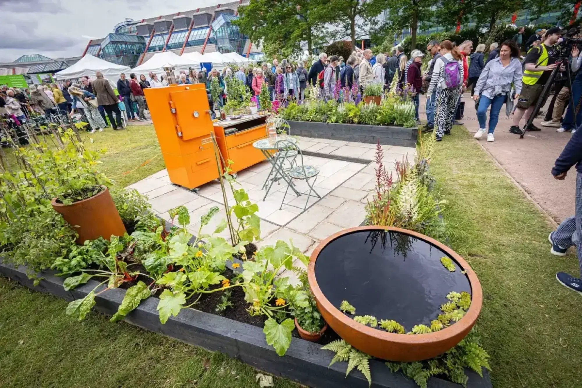 A showcase garden featuring a bright orange outdoor kitchen, a paved patio with a wrought iron bistro set, and raised beds filled with edible plants, flowers, and a large terracotta water feature.
