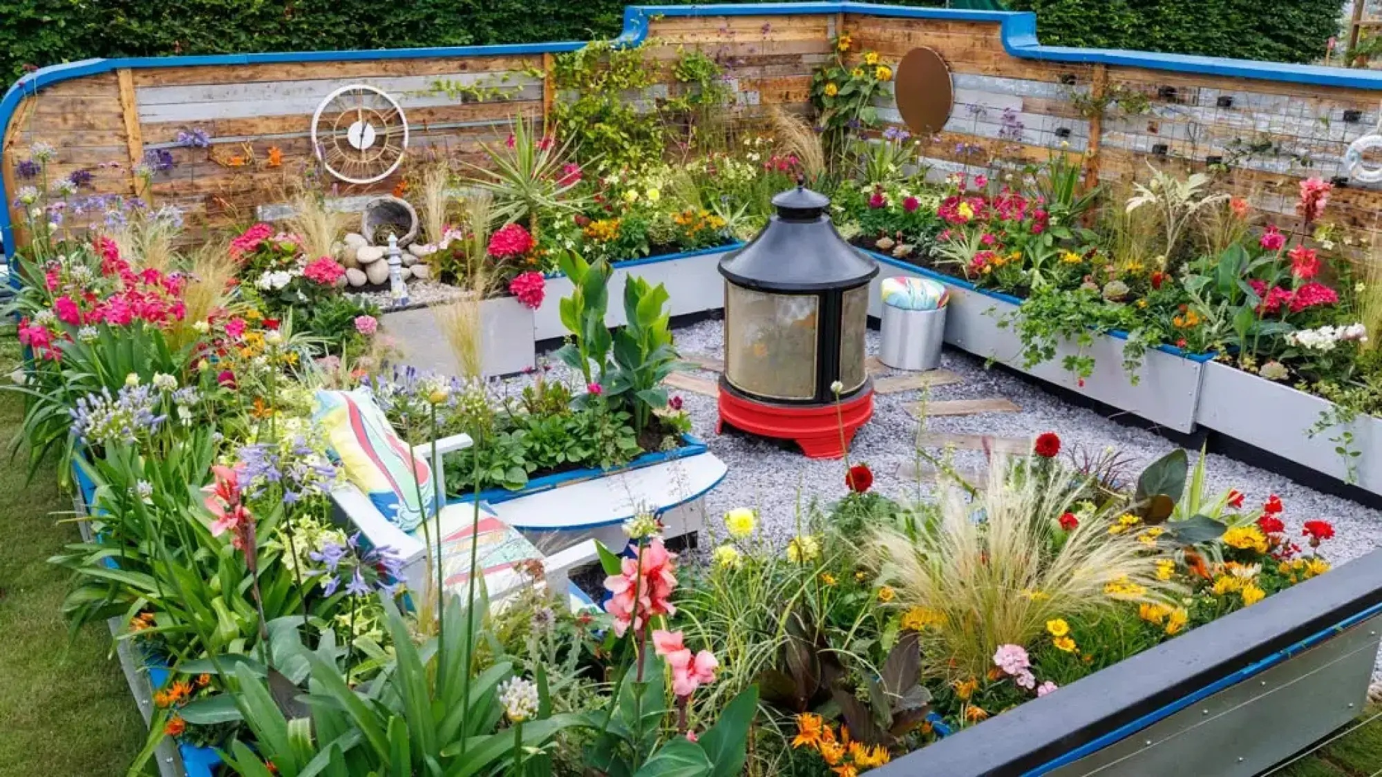 A colourful, nautical-themed garden featuring a black and red lighthouse lantern, boat-shaped flower beds, a reclaimed timber wall with maritime decor, and a white chair.