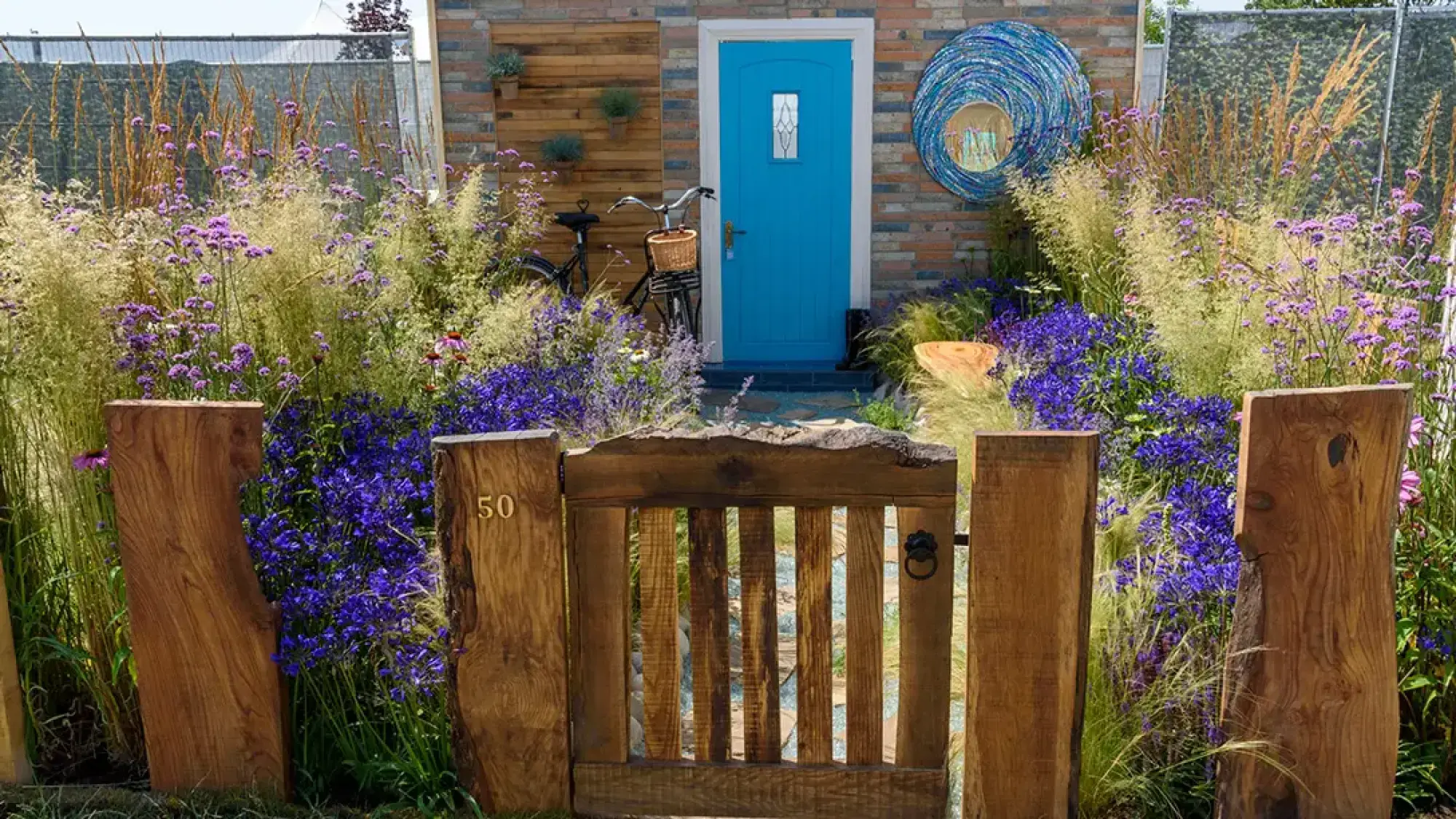 A welcoming greener front garden features a rustic timber gate leading to a bright blue front door, surrounded by abundant purple and blue flowers, and golden ornamental grasses.