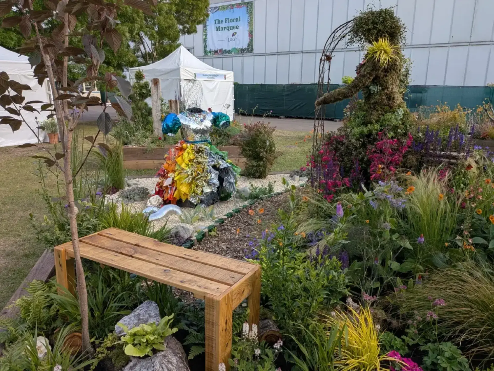 A Beautiful Borders garden features a moss-covered figure, a recycled dress sculpture, and a wooden bench, set in vibrant mixed planting.