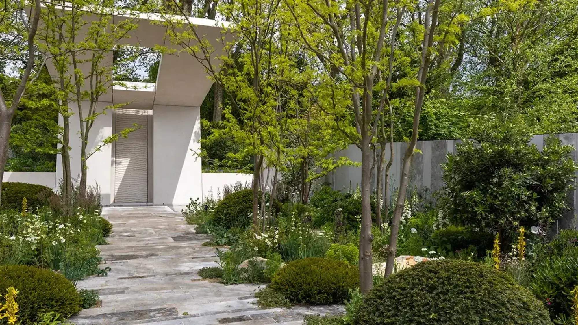 A contemporary show garden with a pale grey archway, textured panel, and a stone path bordered by young trees and lush mixed planting.