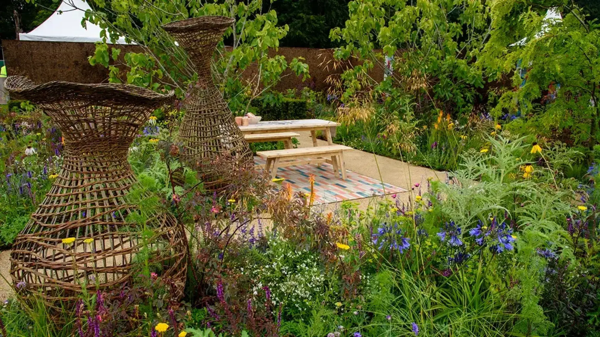 A garden with two large woven willow sculptures, a picnic table on a colourful rug, and lush mixed planting.