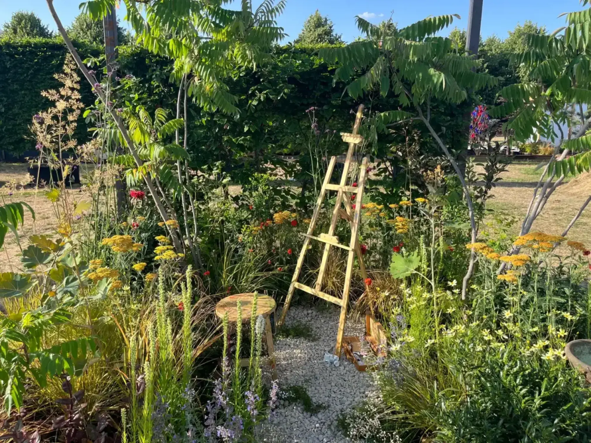 A naturalistic cube garden features a wooden easel and stool on a gravel path, surrounded by vibrant yellow, red, and purple flowers with lush green foliage.