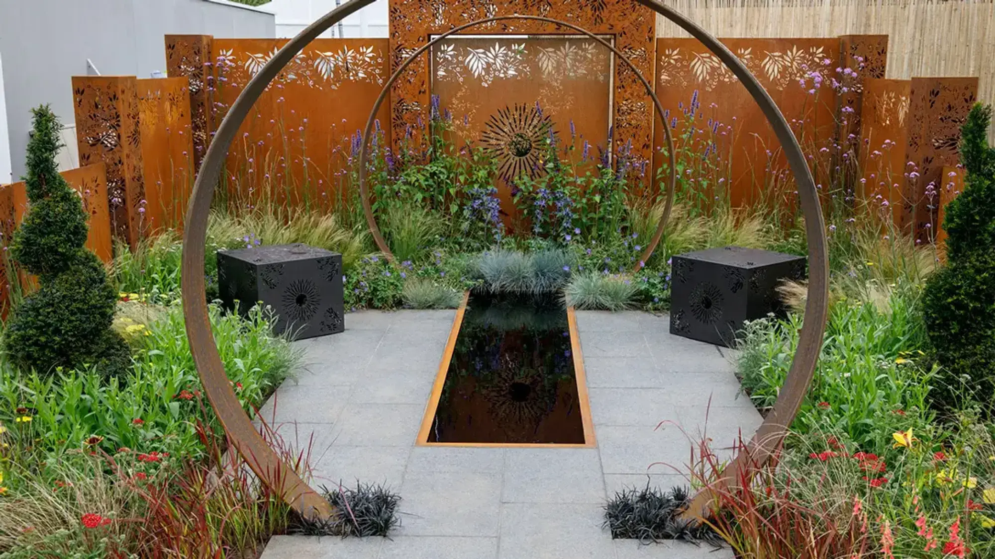 A modern show garden featuring curved Corten steel arches framing a central dark water feature, flanked by black geometric planters and backed by laser-cut screens, amidst grasses and purple flowers.