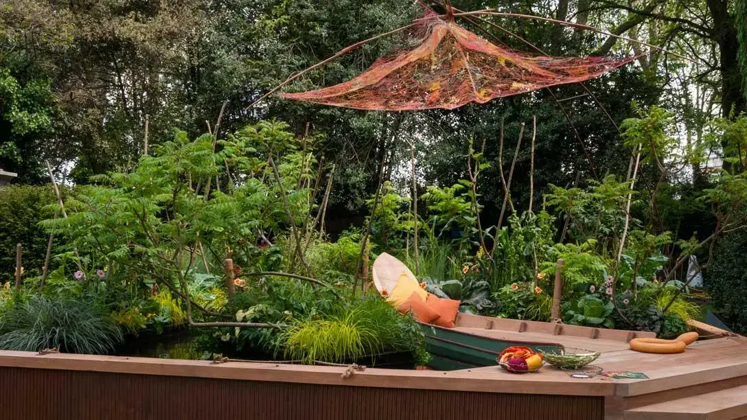 A wooden boat serving as seating floats in a water garden, surrounded by lush green tropical plants, beneath an orange net canopy.