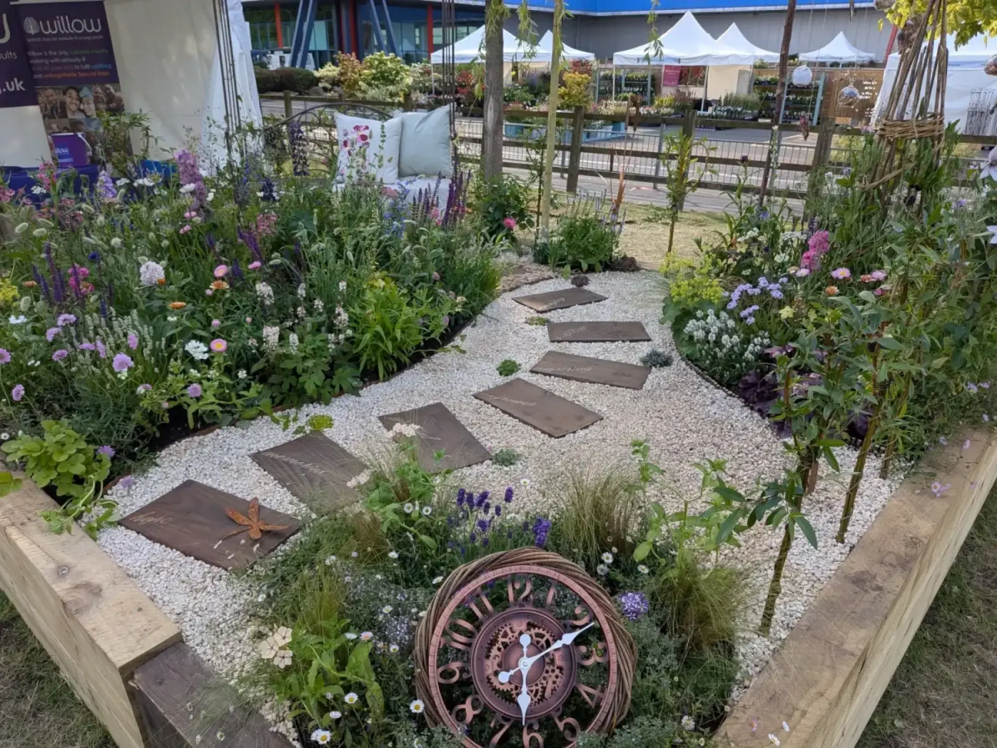 A beautiful border garden features a white gravel path with wooden stepping stones, a woven willow clock sculpture, and informal pink, purple, and white perennial planting surrounding a wrought iron bench.