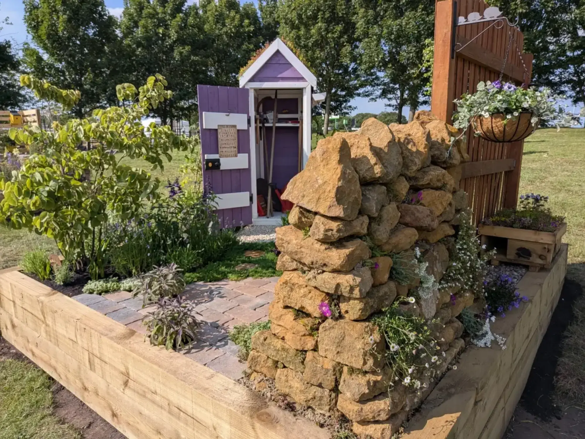 A beautiful border garden features a small purple and white shed, a dry stone wall, and a wooden gate with a floral hanging basket in a raised timber bed.