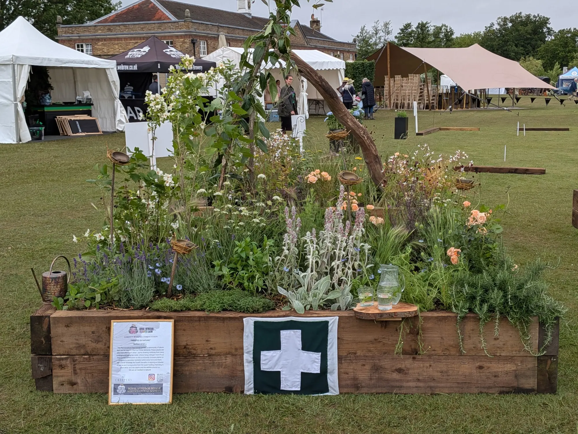 A naturalistic permaculture garden in a rustic timber raised bed, with herbs, flowers, a curved wooden arch, and a white first aid cross.