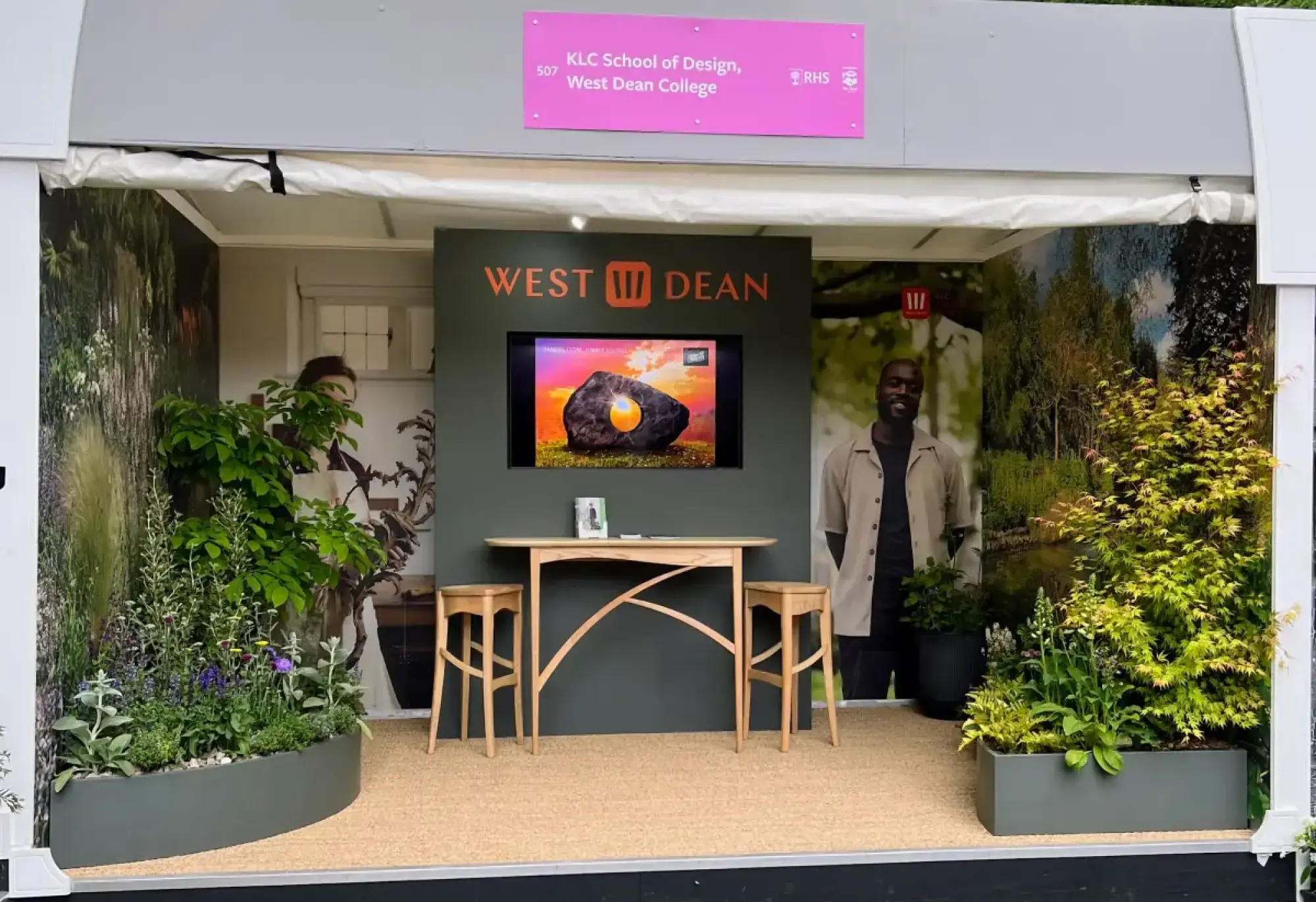 A modern trade stand features a light wooden table and two stools, framed by two grey planters filled with mixed green and purple-flowered plants, all against a dark grey wall with a large screen displaying an abstract rock.