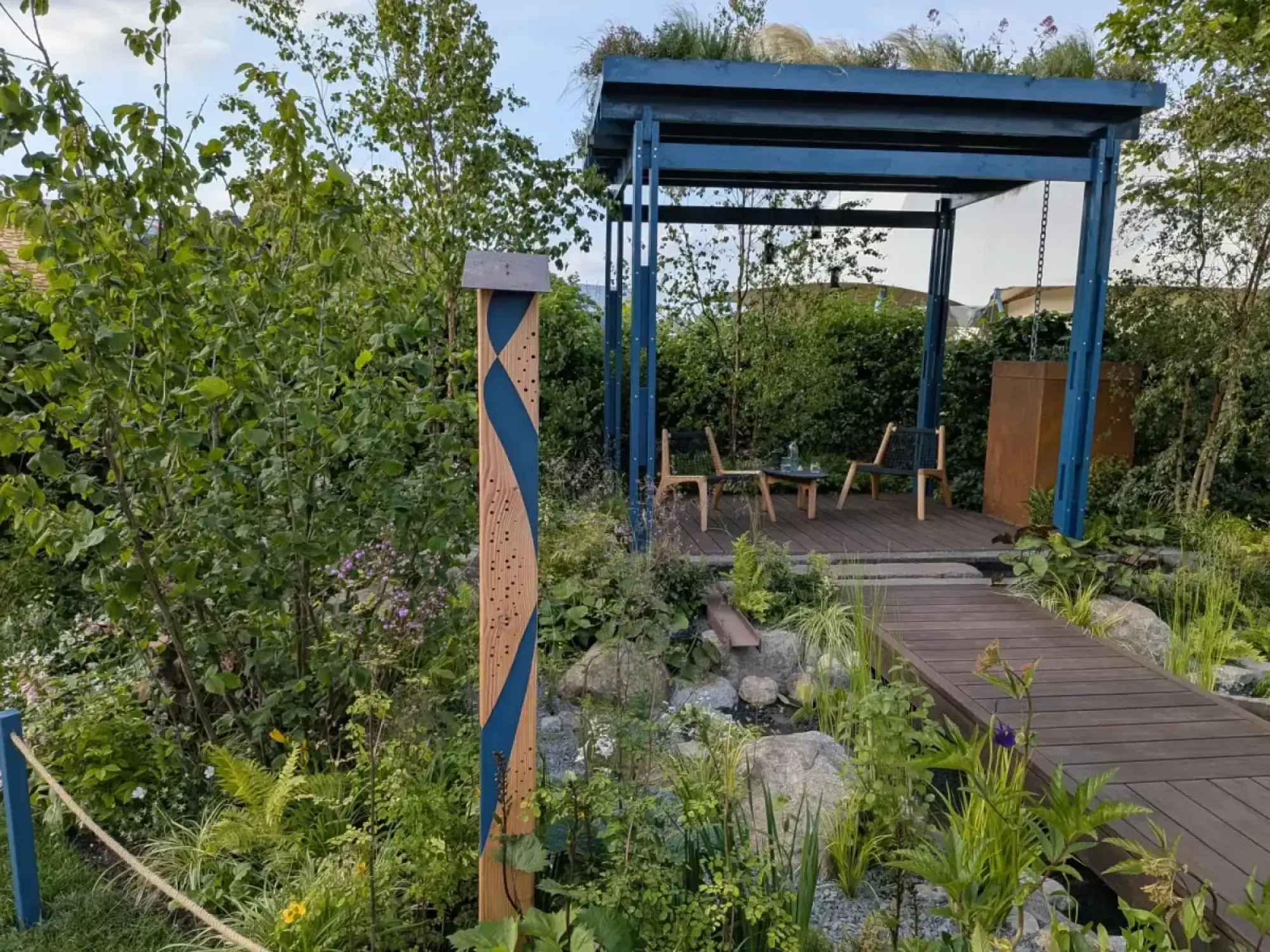 A naturalistic garden features a blue metal pergola with a living roof, a timber boardwalk over a pond, an insect hotel, and lush green planting.