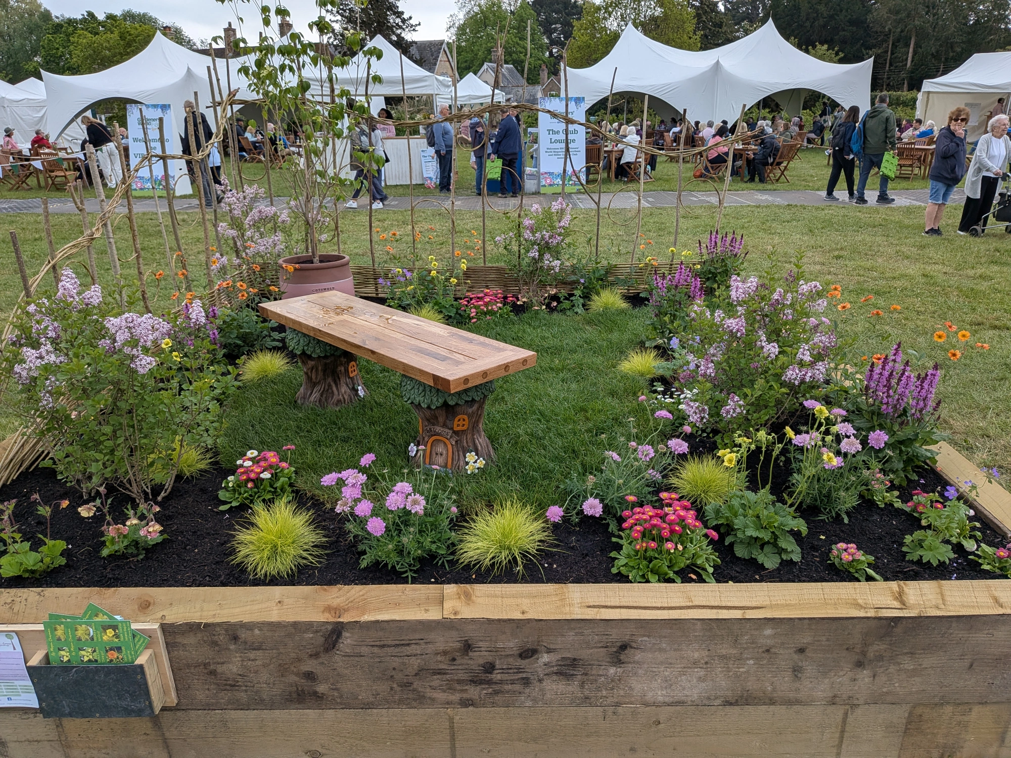 A whimsical show garden with a wooden bench on tree stumps, a willow fence, and borders of purple, pink, and orange flowers.