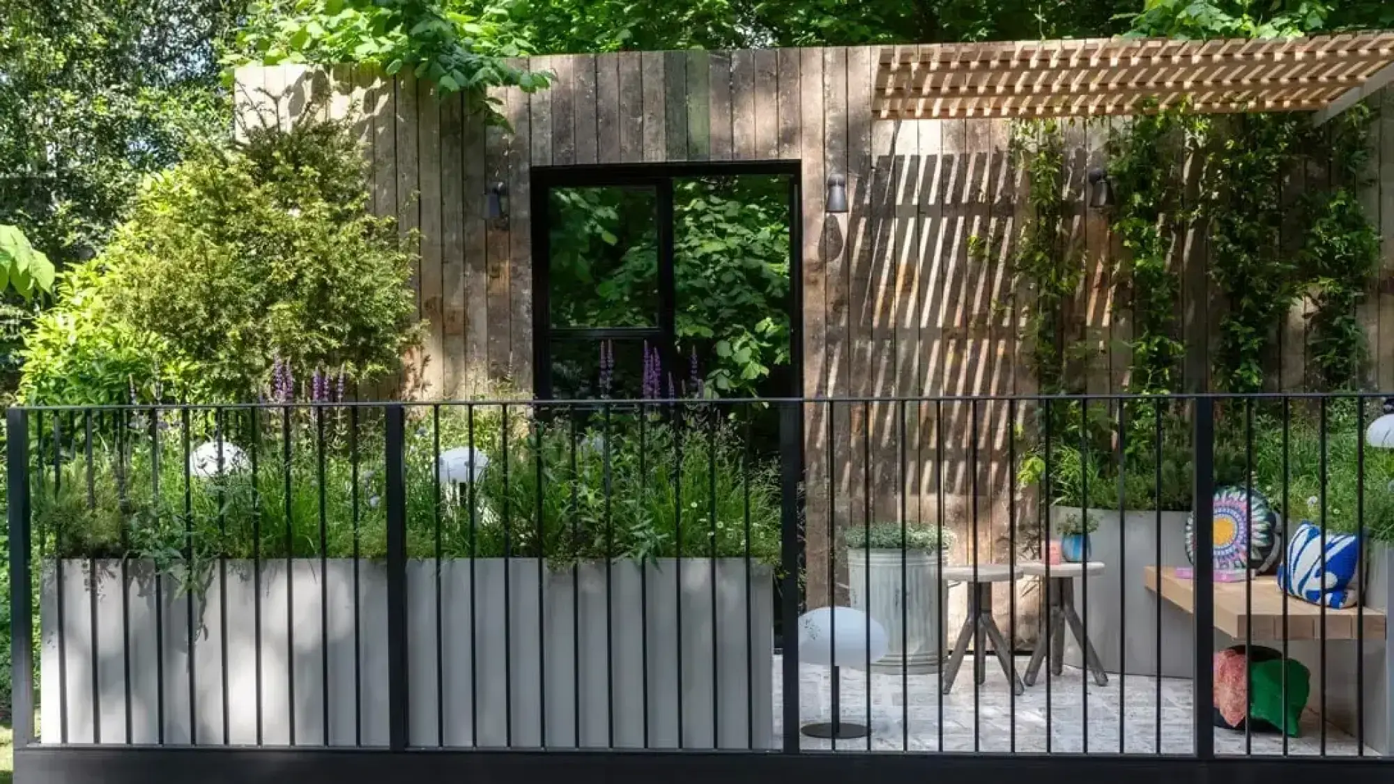 A contemporary balcony garden features a timber wall with a black window, grey planters with greenery, and a wooden bench with colourful cushions.