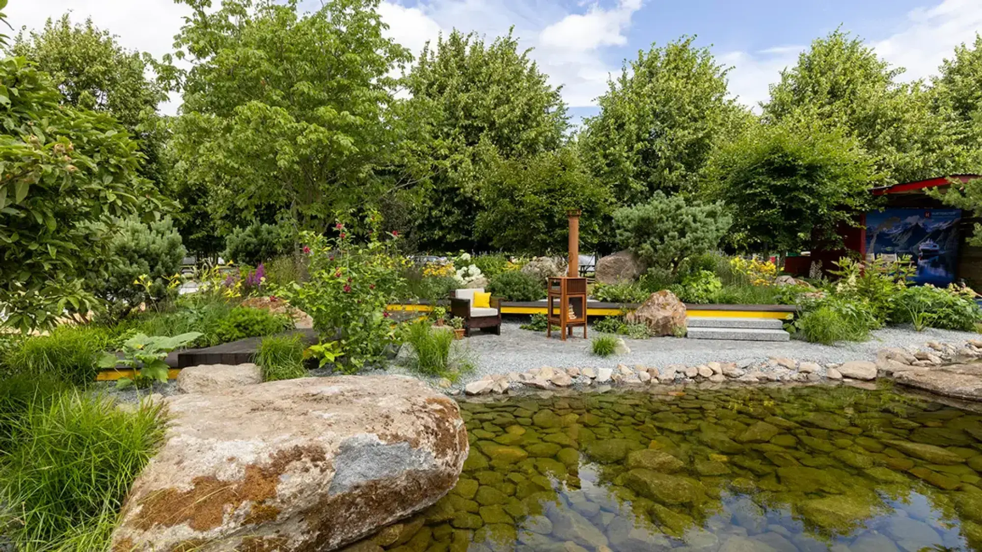 A serene naturalistic show garden features a clear water pond with visible rocks, a grey gravel path, large boulders, and lush mixed perennial planting. A contemporary wooden chair with a yellow cushion and a matching wooden bench with a yellow accent strip sit beside the path, with a tall wooden display structure.