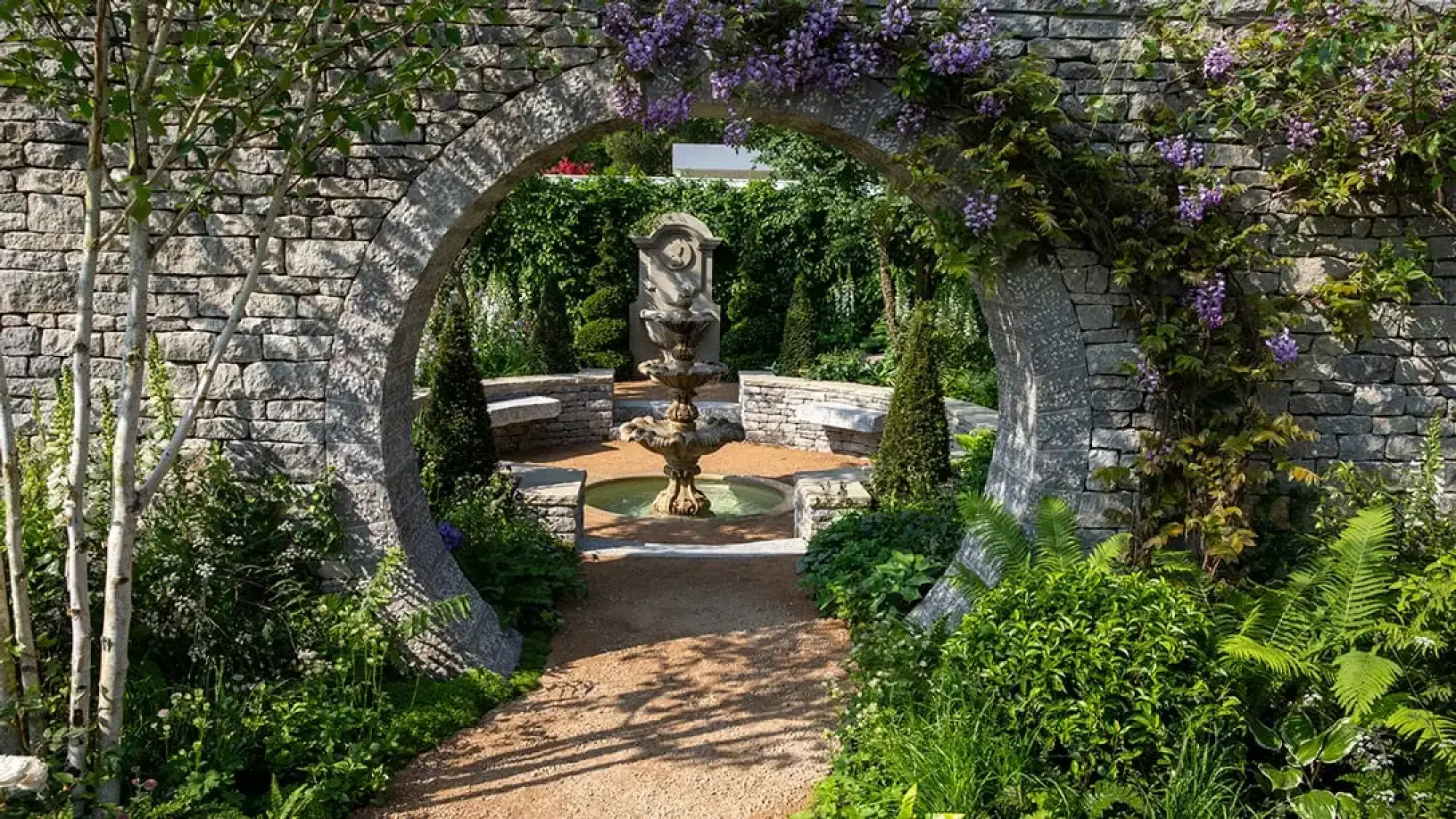 A stone archway draped with purple wisteria opens to a tranquil sanctuary garden featuring a three-tiered fountain, a classical wall fountain, and stone benches.