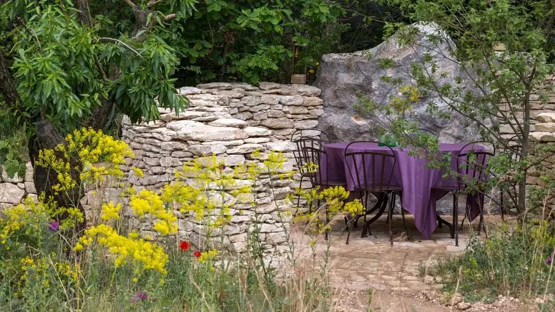 A rustic garden features a dry stone wall, a large boulder, and an outdoor dining set with a purple tablecloth, surrounded by yellow wildflowers and trees.