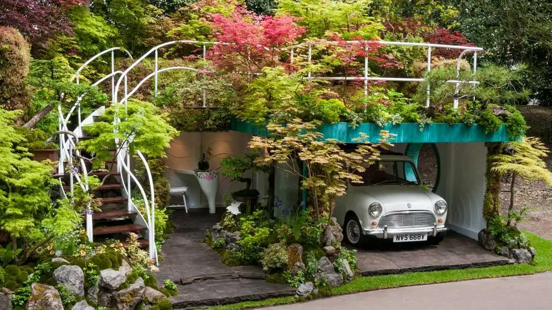 A white Mini car is parked in a teal garage surrounded by a white curved staircase, lush green and red maples, and mossy rocks.