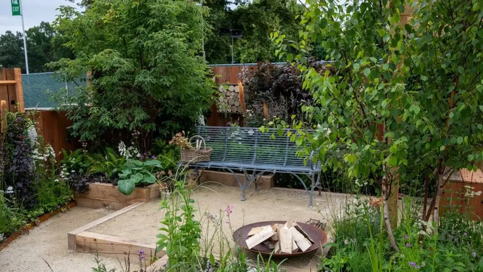 A Get Started Garden with a corten steel fire pit, cast iron bench, gravel, and mixed planting of birch trees, ferns and colourful flowers.