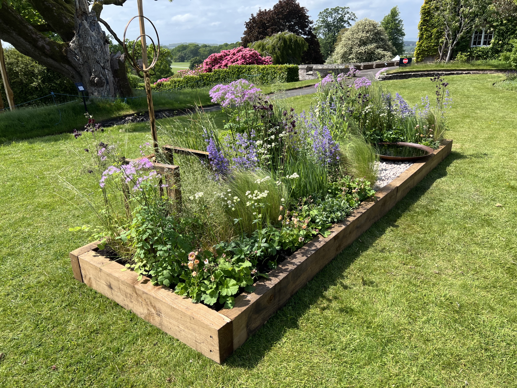 A timber raised bed garden features a rusted metal sculpture, a round water feature, and purple, pink, and white flowers with ornamental grasses.
