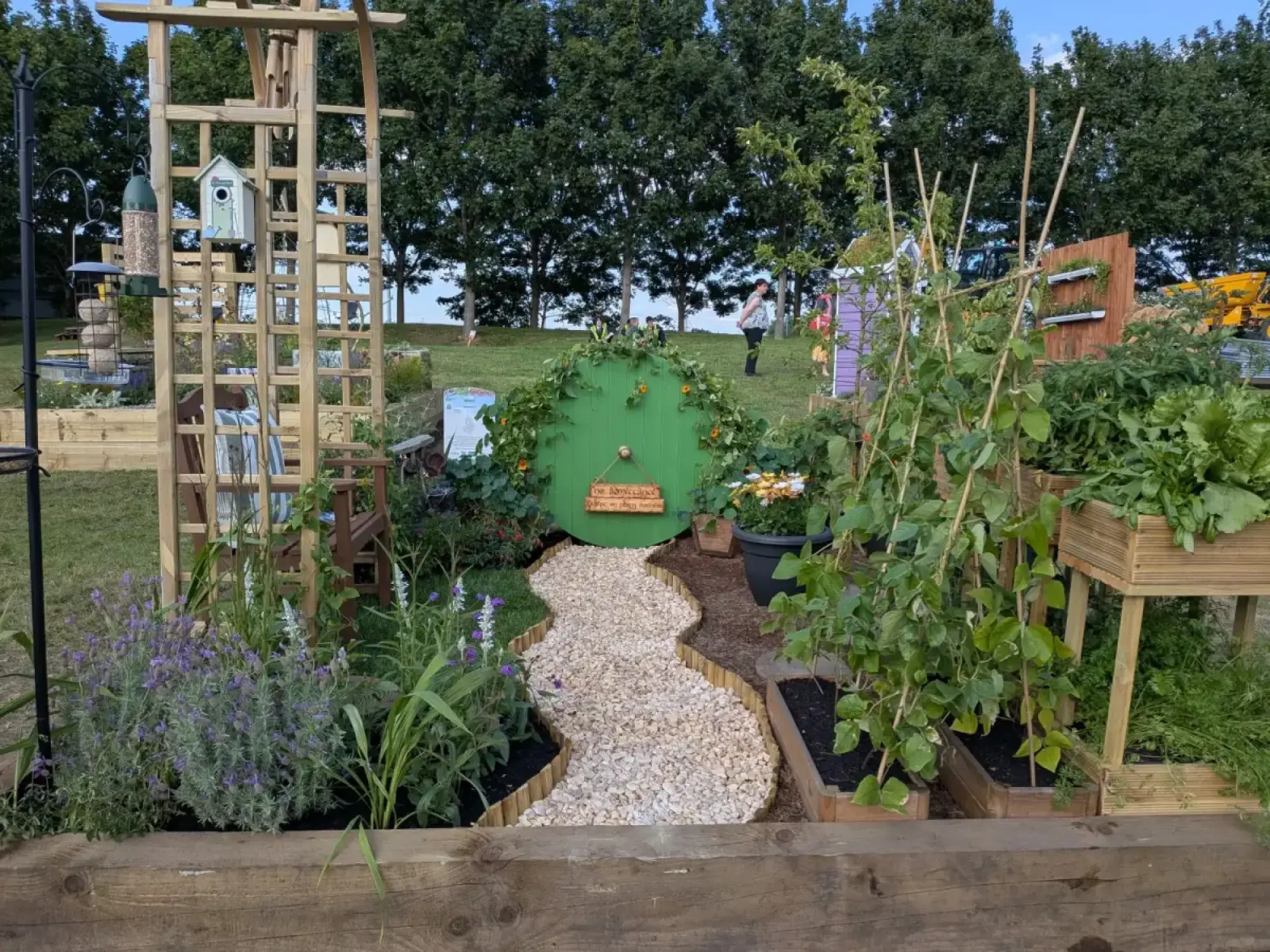 A whimsical garden with a round green hobbit door, a winding gravel path, raised beds of edible plants and lavender, and a wooden arch.