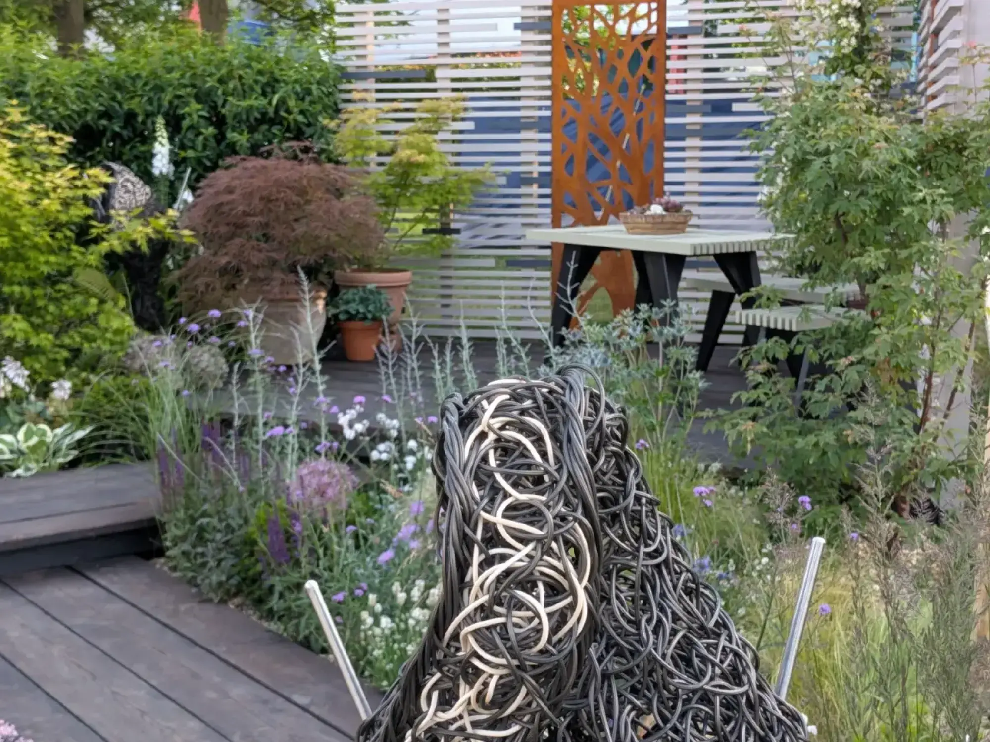 A contemporary garden featuring dark timber decking, a woven sculpture, naturalistic purple and green planting, and a picnic table against a slatted white fence and corten steel screen.