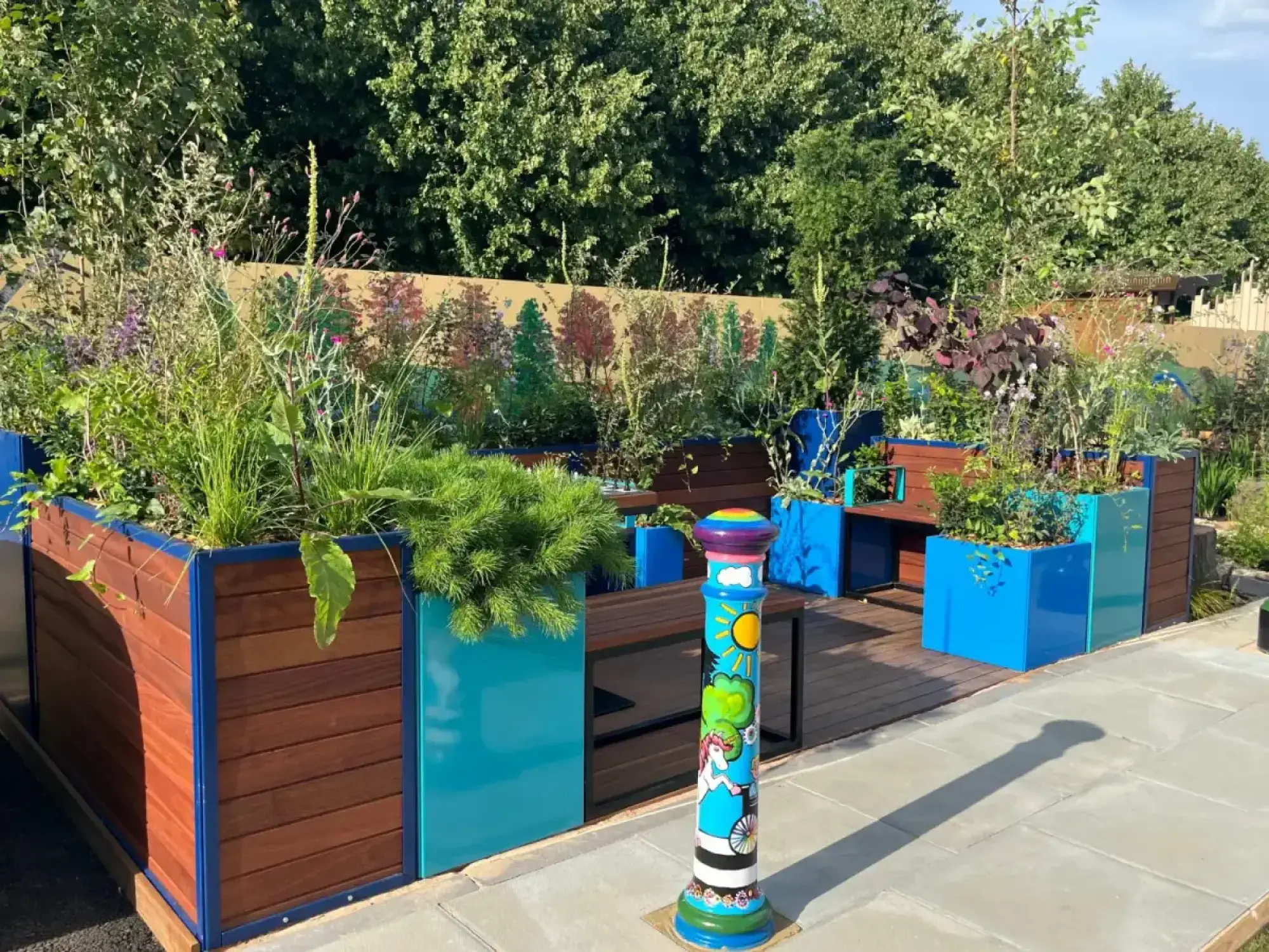 A vibrant urban show garden with timber and blue planters, built-in benches, a wooden deck, mixed planting, grey paving, and a multicoloured bollard.