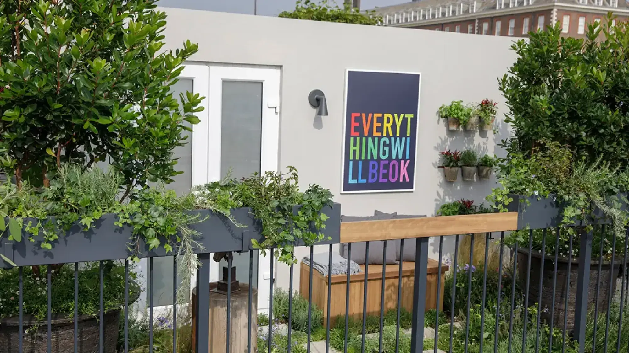 A modern balcony garden features a bright 'EVERYTHING WILL BE OK' sign on a grey wall, vertical herb pots, a wooden storage bench, and lush container planting.