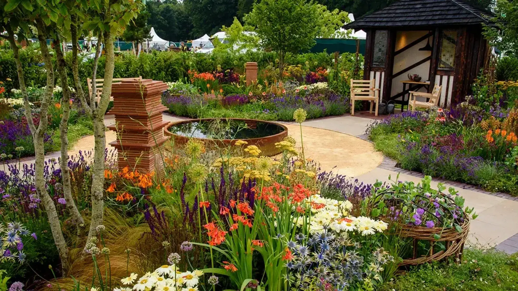 A colourful Arts & Crafts show garden features a rustic wooden gazebo, circular rusted pond, terracotta pillar, and lush mixed perennial borders.