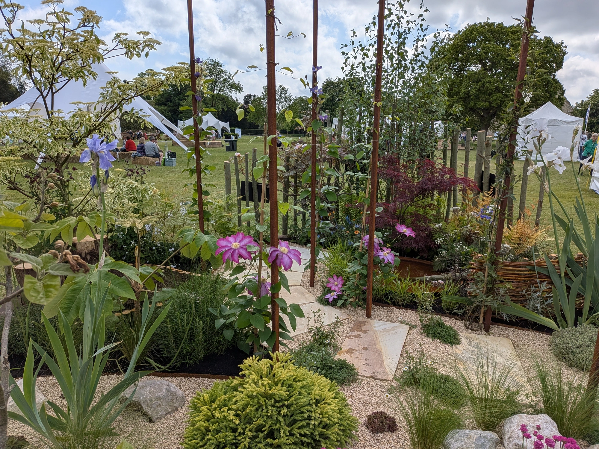 A beautiful border garden features a winding stone path, rust metal posts with purple clematis, a red Japanese maple, and a rusty water bowl.