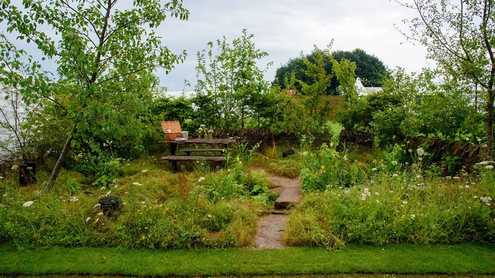 A naturalistic garden displays a rustic wooden picnic bench, dry stone wall, and a winding flagstone path through tall grasses and wildflowers.