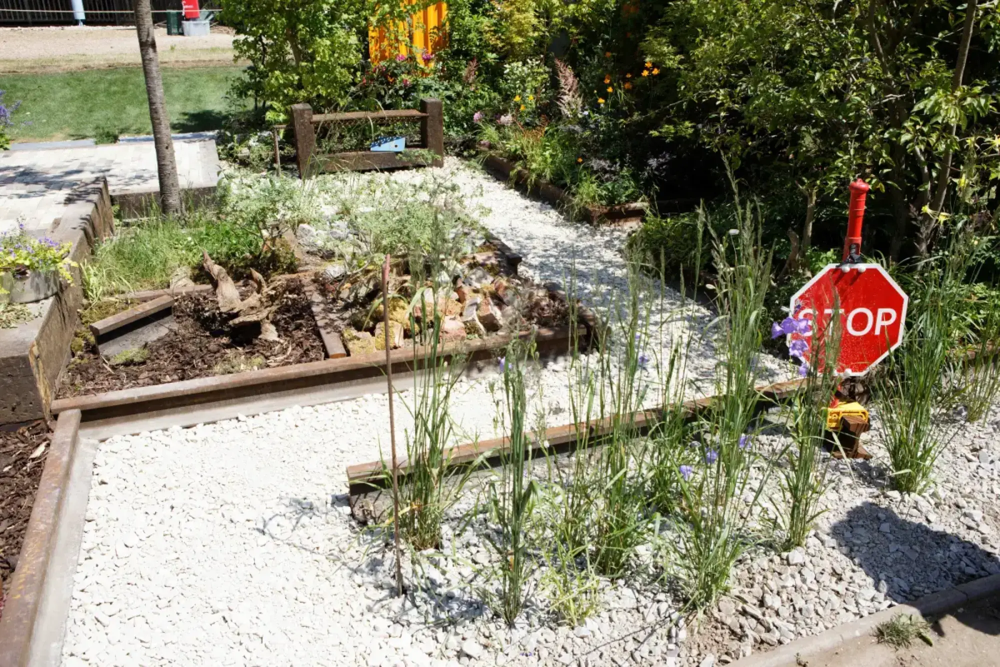A contemporary garden with railway tracks, dark sleepers, white gravel path, uprooted tree stump, red STOP sign, and tall grasses.