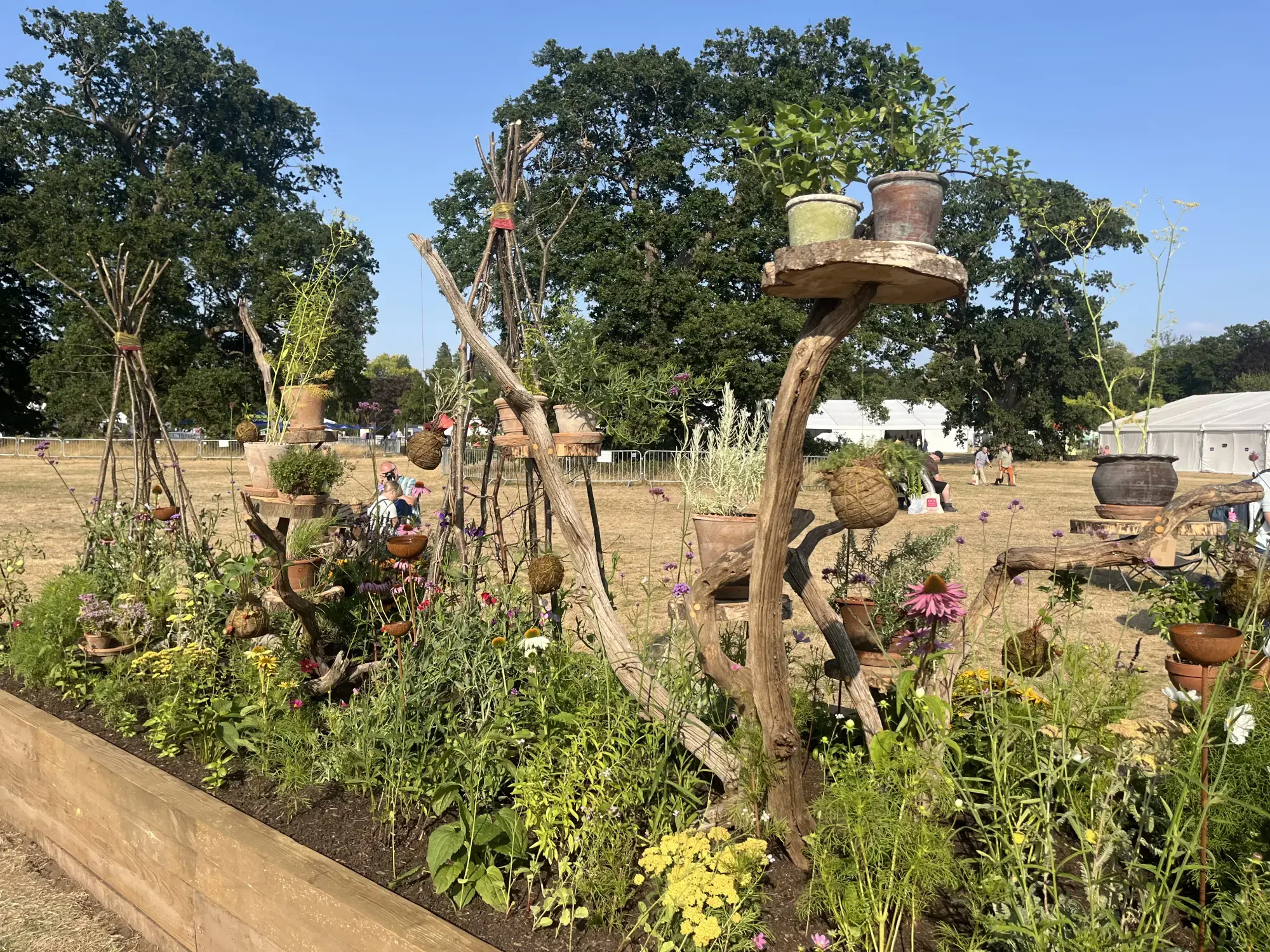 A rustic long border garden in a timber raised bed, with mixed perennials, herbs, and natural timber structures displaying potted plants and kokedama.