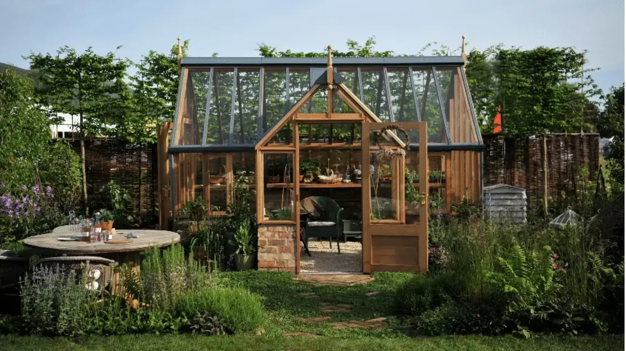 A rustic show garden features a large timber and glass greenhouse, a round wooden dining table, and lush naturalistic planting with herbs and ferns.